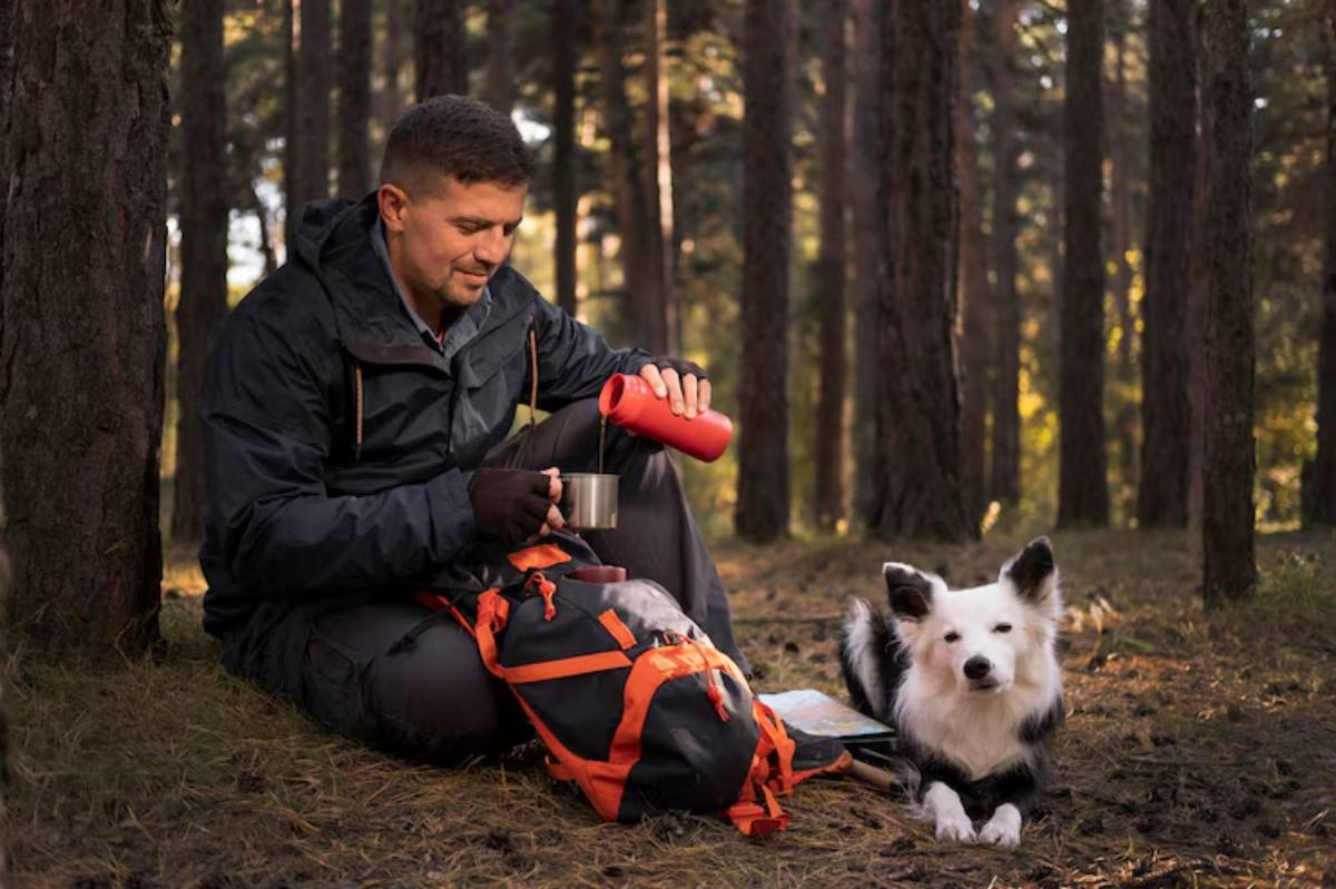 A person sits on the forest floor, pouring a drink from a red thermos, while a fluffy black and white dog lounges nearby. 