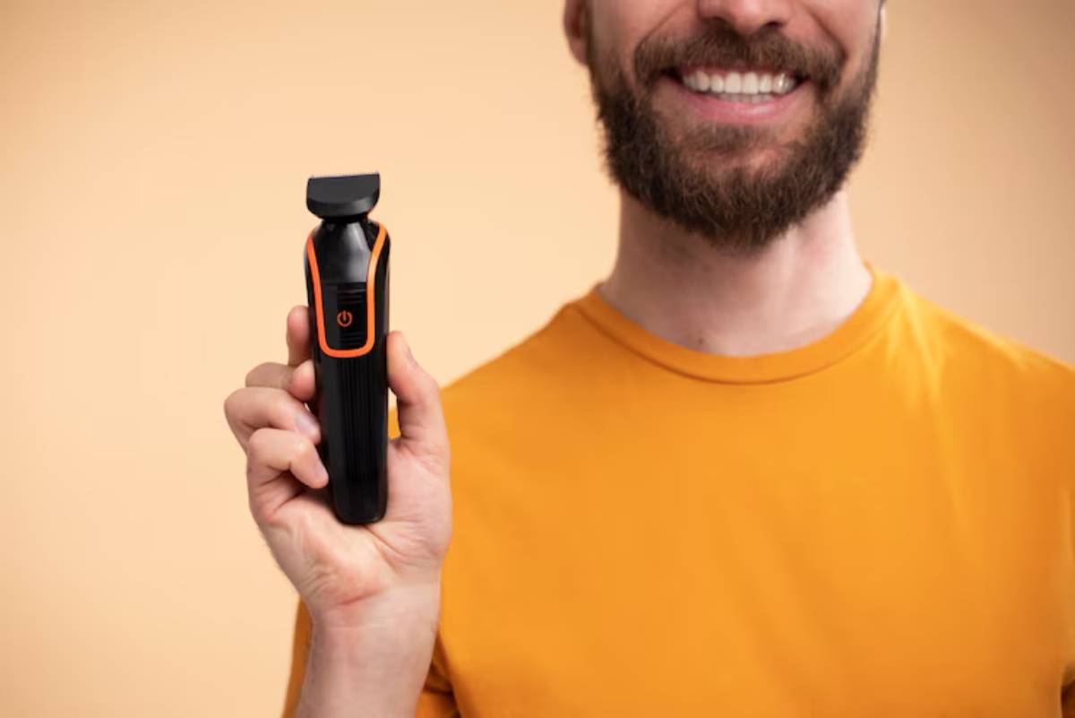  A man in an orange shirt smiles while holding a black and orange trimmer against a light orange background.