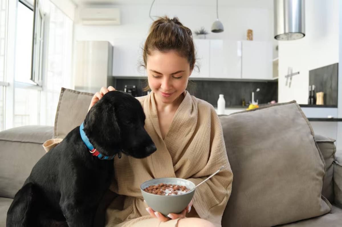 A woman in a bathrobe sits on a couch, holding a bowl of cereal, while a black dog curiously leans in beside her. 